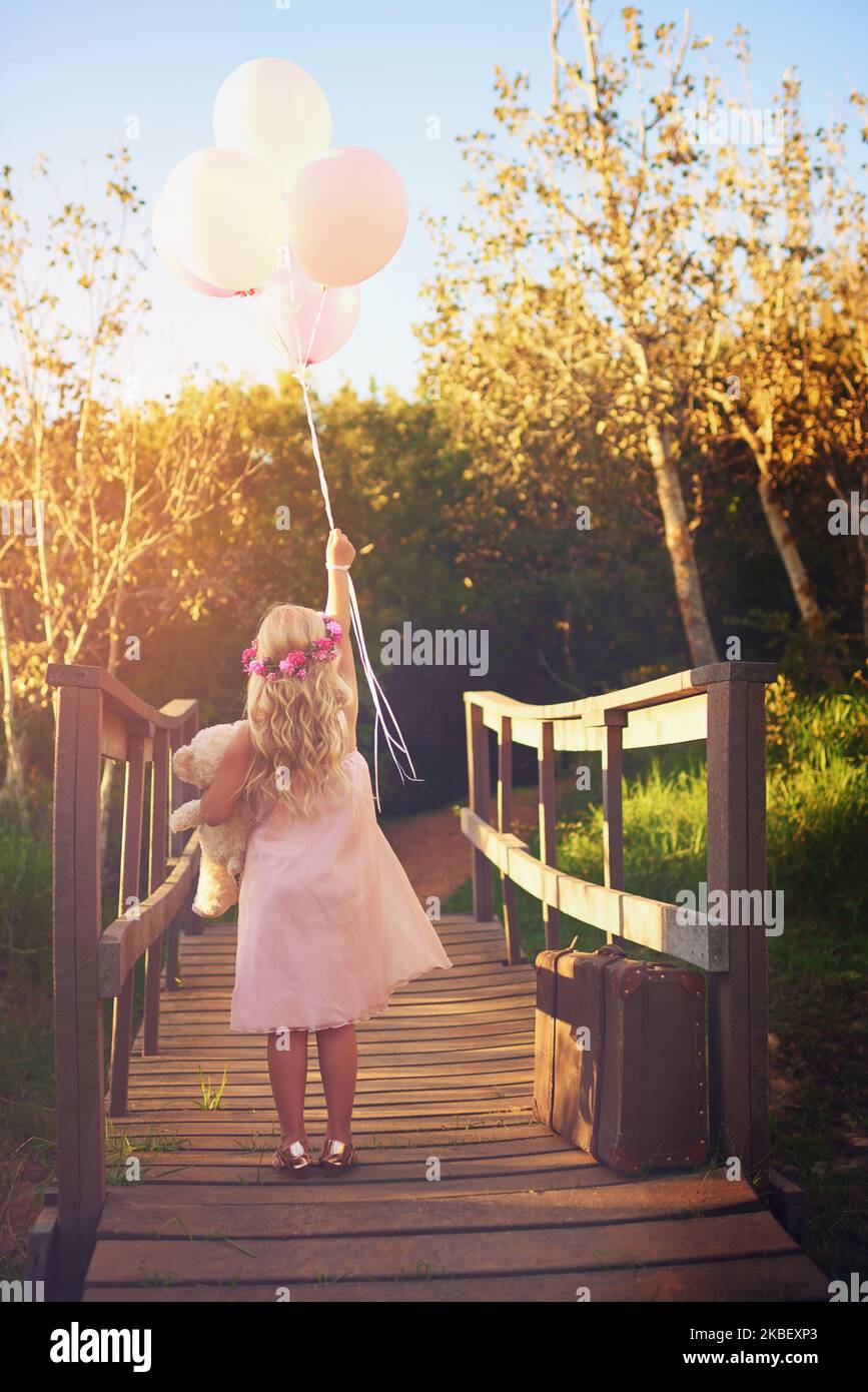 Auf, auf und weg. Ein unkenntlich kleines Mädchen, das Luftballons und einen Teddybären in der Mitte einer Brücke hält. Stockfoto