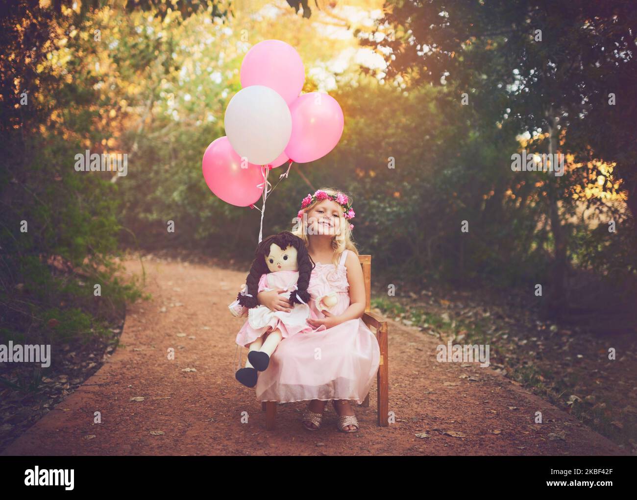 Ich kann hier den ganzen Tag sitzen. Ein glückliches kleines Mädchen sitzt und wartet mit einer Puppe und Luftballons mitten auf einer unbefestigten Straße. Stockfoto