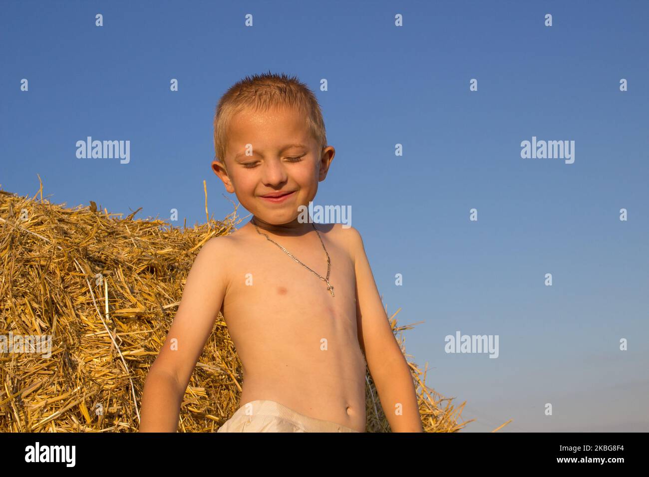 Ein kleiner Junge auf einem Feld in der Nähe eines Strohhalms schloss seine Augen vor dem Sonnenuntergang Stockfoto