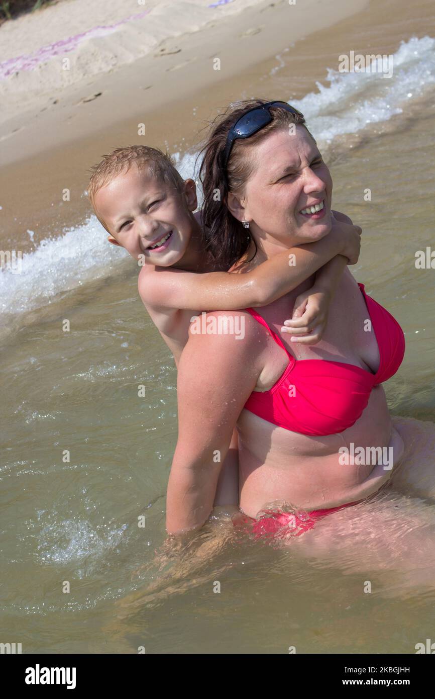 Der Sohn umarmt den Hals seiner Mutter im Meer hinter ihm im Wasser Stockfoto