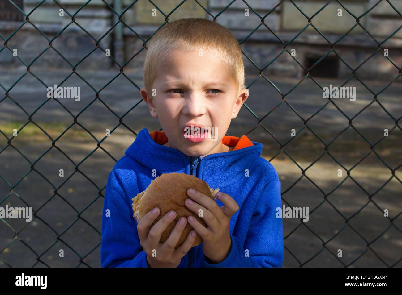 Der Junge hebt sein Gesicht vom Chezburger auf dem Hintergrund des Volleyballzauns Stockfoto