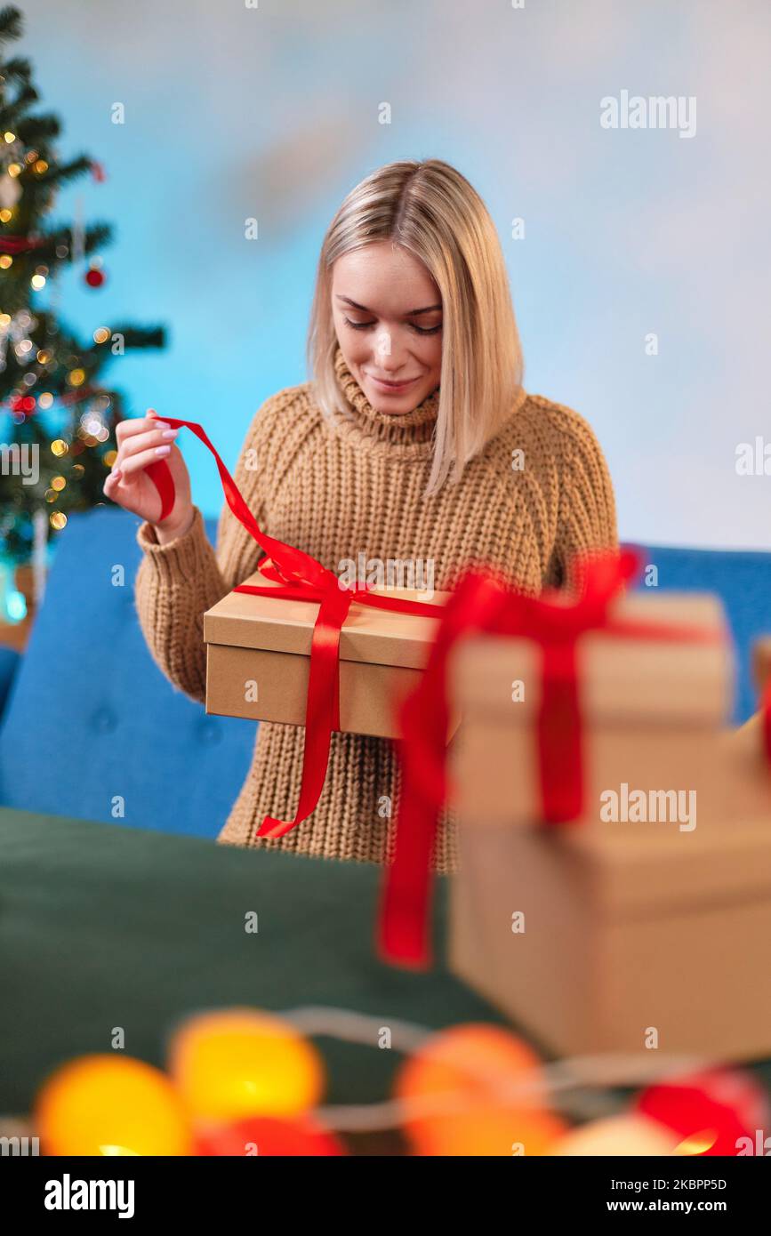 Eine Frau in einem Pullover feiert das neue Jahr und öffnet ein Geschenk, das unter dem Baum gefunden wird. Urlaubskonzept. Das Mädchen packt ein Weihnachtsgeschenk aus. Nahaufnahme Stockfoto