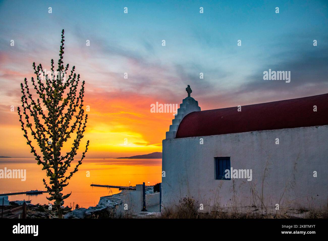 Verträumter Sonnenuntergang während der goldenen Stunde mit einer kleinen traditionellen Kapelle kurz vor der Dämmerung mit dem Himmel in Rot, Orange und warmen Farben, wenn die Sonne hinter der Ägäis über Mykonos Stadt oder Chora untergeht, auf Myconos Insel in den Kykladen, Ägäis in Griechenland. Die berühmte griechische Mittelmeerinsel wird mit weiß getünchten traditionellen Gebäuden wie Windmühlen oder einer kleinen Kirche als Insel der Winde bezeichnet. Mykonos ist eine beliebte Insel für Prominente und Touristen, die im pulsierenden Nachtleben feiern möchten. Die Insel gilt auch als homosexuell freundlich. Die griechische Regierung hat den Sommertouristen wieder belebt Stockfoto