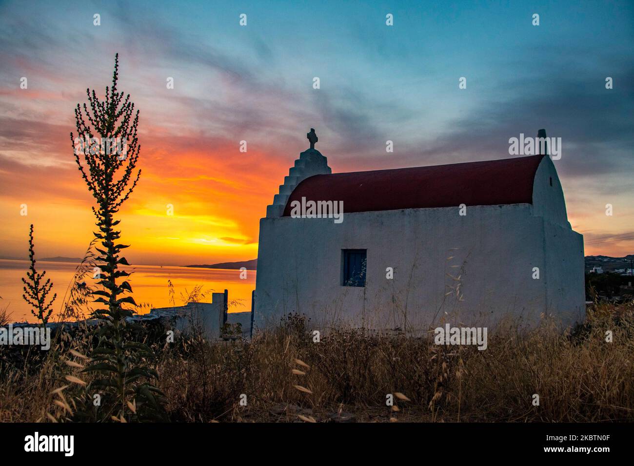 Verträumter Sonnenuntergang während der goldenen Stunde mit einer kleinen traditionellen Kapelle kurz vor der Dämmerung mit dem Himmel in Rot, Orange und warmen Farben, wenn die Sonne hinter der Ägäis über Mykonos Stadt oder Chora untergeht, auf Myconos Insel in den Kykladen, Ägäis in Griechenland. Die berühmte griechische Mittelmeerinsel wird mit weiß getünchten traditionellen Gebäuden wie Windmühlen oder einer kleinen Kirche als Insel der Winde bezeichnet. Mykonos ist eine beliebte Insel für Prominente und Touristen, die im pulsierenden Nachtleben feiern möchten. Die Insel gilt auch als homosexuell freundlich. Die griechische Regierung hat den Sommertouristen wieder belebt Stockfoto