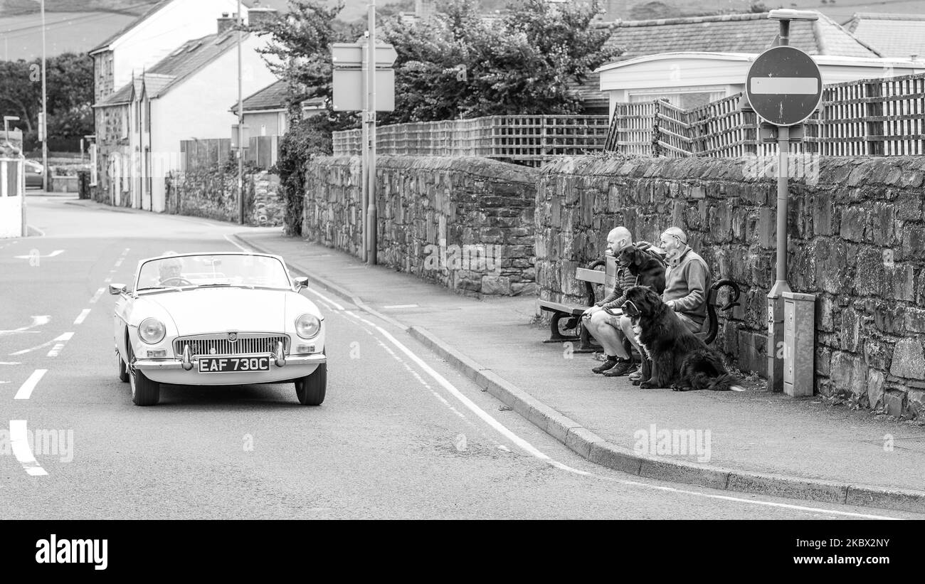 Eine Schwarz-Weiß-Aufnahme eines Oldtimers, der an Menschen vorbeifährt, die auf einer Bank sitzen Stockfoto