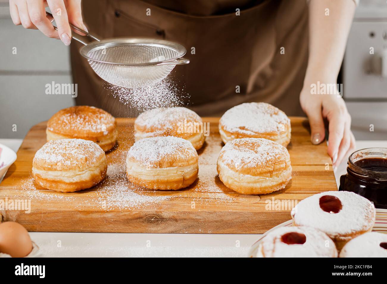 Frau bereitet frische Donuts mit Marmelade in der heimischen Küche. Kochen traditionelle jüdische Hanukkah sufganiyot. Hände bestreuen Berliner mit Puderzucker. Stockfoto