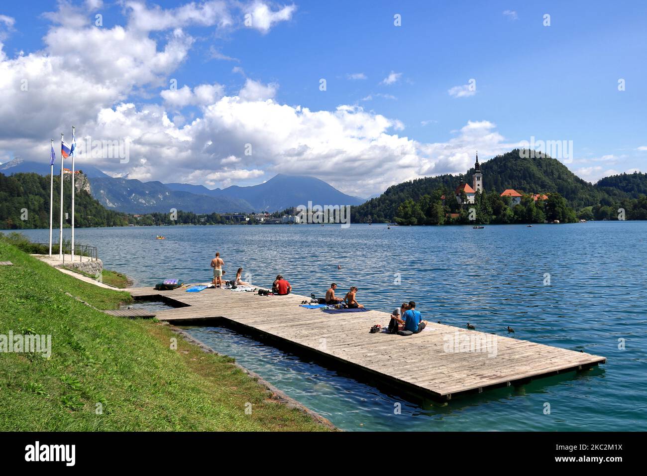 Die Menschen, die auf einem schwimmenden Dock am Bleder See in Slowenien mit der Mariä Himmelfahrt Kirche vor ihnen sitzen Stockfoto