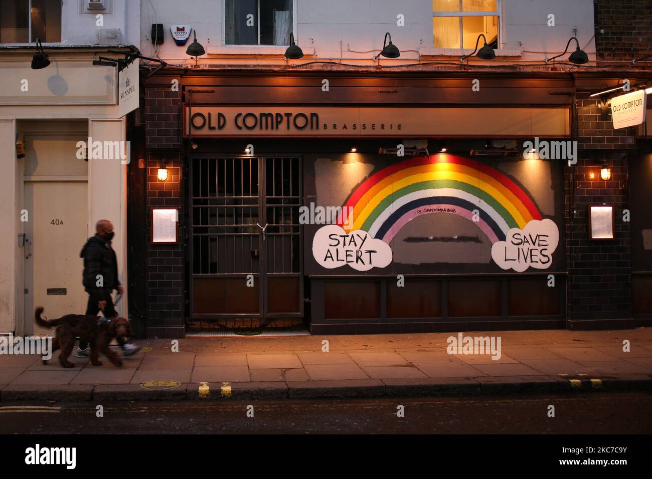 Ein Mann mit einer Gesichtsmaske geht an einem Regenbogengemälde vorbei, das die Menschen auffordert, „Stay Alert, Save Lives“ zu sehen, das am 12. Januar 2021 auf dem Boarding einer vorübergehend geschlossenen Brasserie in einer nahe verlassenen Old Compton Street im Londoner Stadtteil Soho gemalt wurde. England begann heute die zweite volle Woche seiner dritten nationalen Coronavirus-Sperre, die vom britischen Premierminister Boris Johnson aufgrund der Warnungen der Gesundheitsbehörden verhängt wurde, dass der National Health Service (NHS) bald mit Patienten mit Covid-19 überfordert werden könnte. (Foto von David Cliff/NurPhoto) Stockfoto