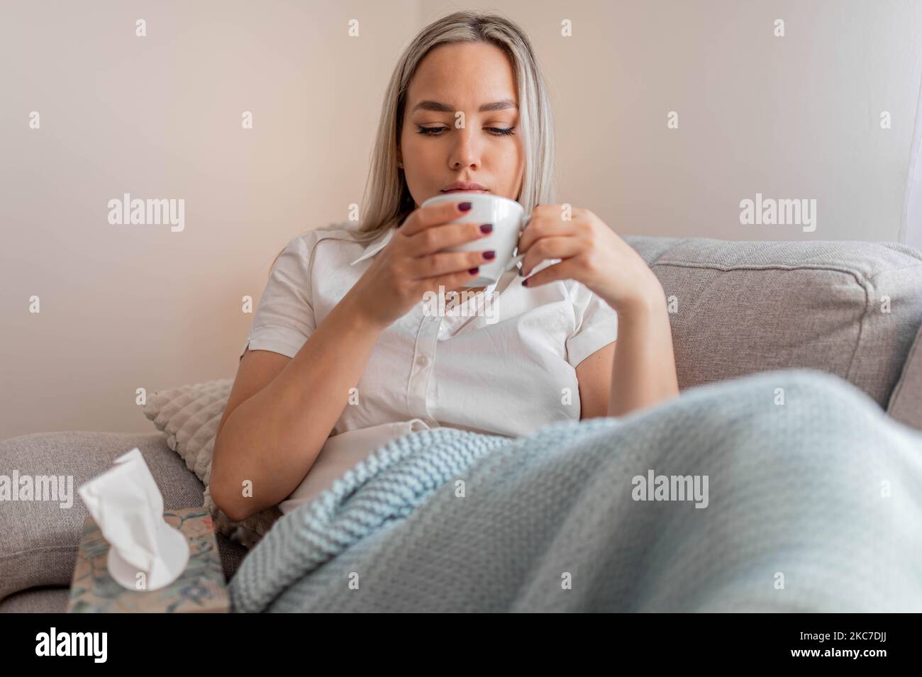 Kranke Frau, die unter der Decke sitzt. Kranke Frau mit saisonalen Infektionen, Grippe, Allergie im Bett liegend. Kranke Frau mit einer Decke bedeckt, die Tee trinkt Stockfoto