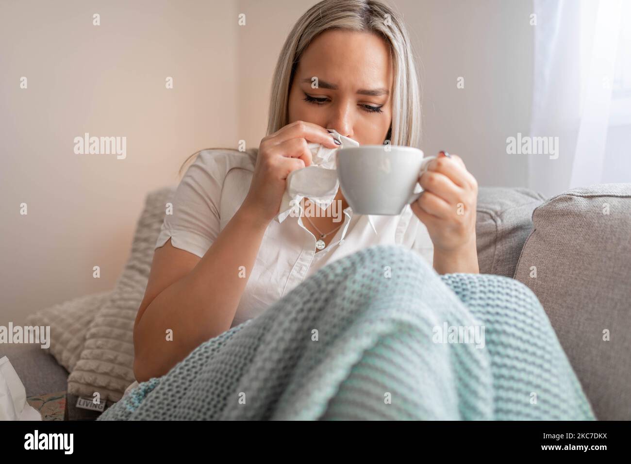 Kranke Frau, die unter der Decke sitzt. Kranke Frau mit saisonalen Infektionen, Grippe, Allergie im Bett liegend. Kranke Frau mit einer Decke bedeckt, die Tee trinkt Stockfoto