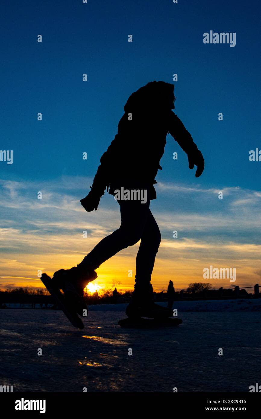 Dunkle Silhouetten von Menschen beim Eislaufen auf dem kristallklaren natürlichen gefrorenen See in der Region Nordbrabant am Nachmittag vor dem Sonnenuntergang. Eislaufrausch übernimmt die Niederlande. Die Niederländer haben viel Spaß und genießen das einwöchige, kalte Schneewetter mit Temperaturen unter Null, die dazu führen, dass Seen, Bäche, Teiche, Flüsse und Kanäle gefrieren. Im Park Meerland in der Nähe der Stadt Eindhoven haben sich die Massen an einem sonnigen Tag mit blauem Himmel abgesetzt, da es eine niederländische Tradition ist. Außerdem werden die Menschen im Freien in der eisgefrorenen Natur und der kalten Umgebung gesehen, die auf dem Eis der gefrorenen Seen spazieren gehen, p Stockfoto