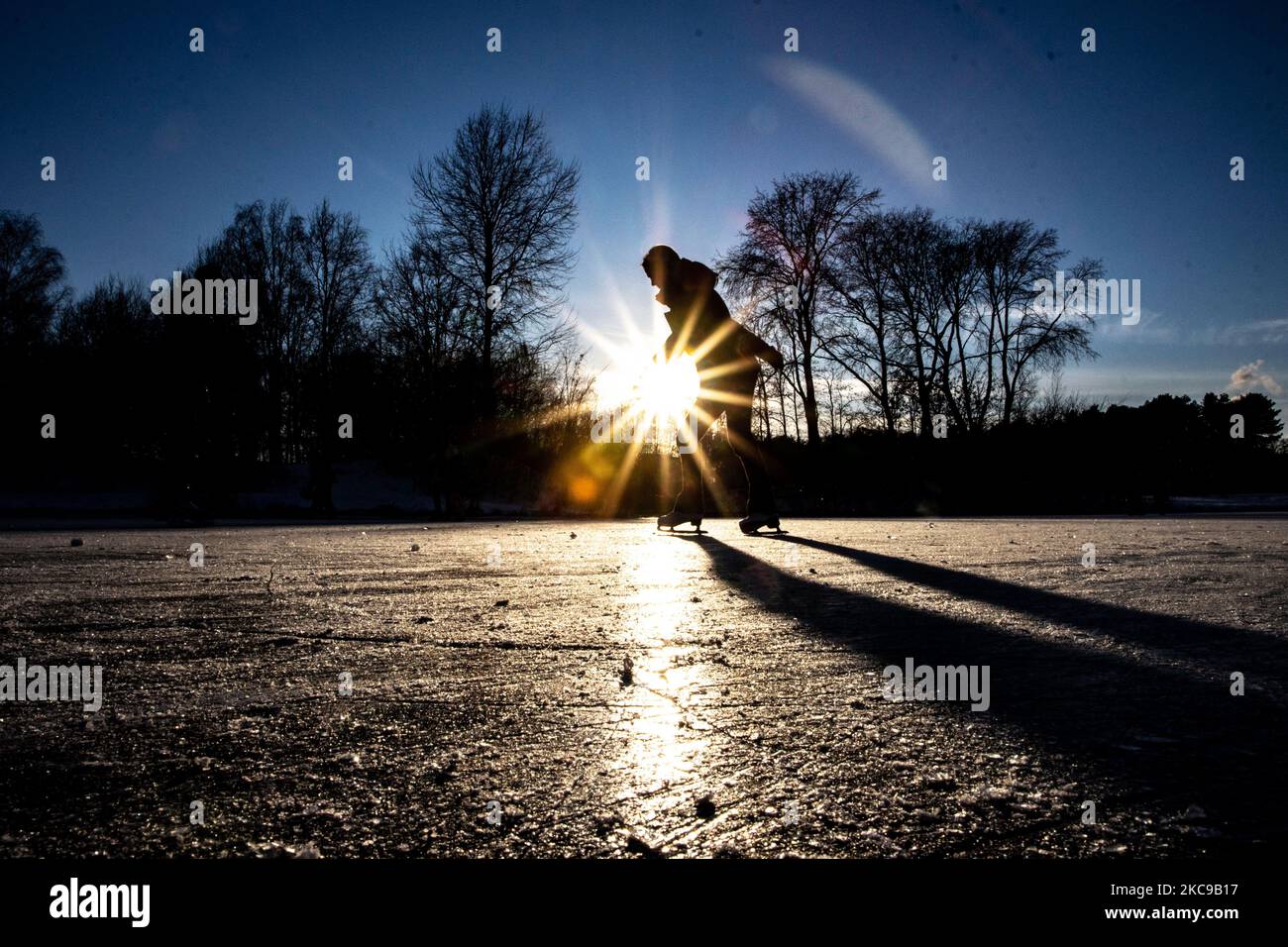 Dunkle Silhouetten von Menschen beim Eislaufen auf dem kristallklaren natürlichen gefrorenen See in der Region Nordbrabant am Nachmittag vor dem Sonnenuntergang. Eislaufrausch übernimmt die Niederlande. Die Niederländer haben viel Spaß und genießen das einwöchige, kalte Schneewetter mit Temperaturen unter Null, die dazu führen, dass Seen, Bäche, Teiche, Flüsse und Kanäle gefrieren. Im Park Meerland in der Nähe der Stadt Eindhoven haben sich die Massen an einem sonnigen Tag mit blauem Himmel abgesetzt, da es eine niederländische Tradition ist. Außerdem werden die Menschen im Freien in der eisgefrorenen Natur und der kalten Umgebung gesehen, die auf dem Eis der gefrorenen Seen spazieren gehen, p Stockfoto