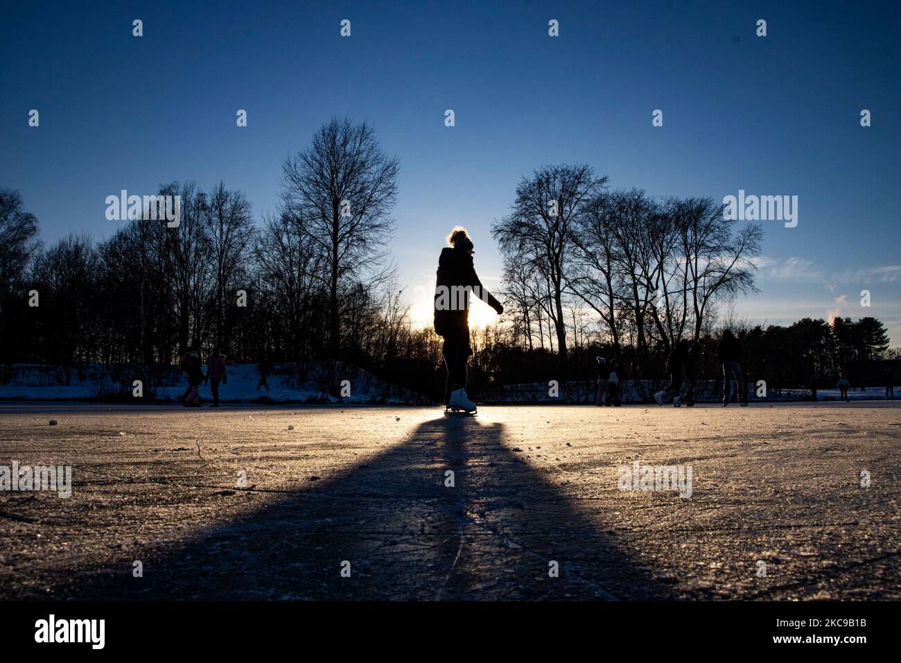 Dunkle Silhouetten von Menschen beim Eislaufen auf dem kristallklaren natürlichen gefrorenen See in der Region Nordbrabant am Nachmittag vor dem Sonnenuntergang. Eislaufrausch übernimmt die Niederlande. Die Niederländer haben viel Spaß und genießen das einwöchige, kalte Schneewetter mit Temperaturen unter Null, die dazu führen, dass Seen, Bäche, Teiche, Flüsse und Kanäle gefrieren. Im Park Meerland in der Nähe der Stadt Eindhoven haben sich die Massen an einem sonnigen Tag mit blauem Himmel abgesetzt, da es eine niederländische Tradition ist. Außerdem werden die Menschen im Freien in der eisgefrorenen Natur und der kalten Umgebung gesehen, die auf dem Eis der gefrorenen Seen spazieren gehen, p Stockfoto