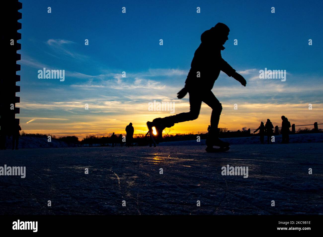 Dunkle Silhouetten von Menschen beim Eislaufen auf dem kristallklaren natürlichen gefrorenen See in der Region Nordbrabant am Nachmittag vor dem Sonnenuntergang. Eislaufrausch übernimmt die Niederlande. Die Niederländer haben viel Spaß und genießen das einwöchige, kalte Schneewetter mit Temperaturen unter Null, die dazu führen, dass Seen, Bäche, Teiche, Flüsse und Kanäle gefrieren. Im Park Meerland in der Nähe der Stadt Eindhoven haben sich die Massen an einem sonnigen Tag mit blauem Himmel abgesetzt, da es eine niederländische Tradition ist. Außerdem werden die Menschen im Freien in der eisgefrorenen Natur und der kalten Umgebung gesehen, die auf dem Eis der gefrorenen Seen spazieren gehen, p Stockfoto