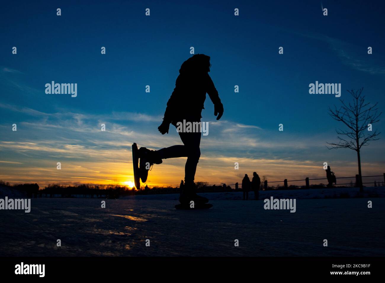 Dunkle Silhouetten von Menschen beim Eislaufen auf dem kristallklaren natürlichen gefrorenen See in der Region Nordbrabant am Nachmittag vor dem Sonnenuntergang. Eislaufrausch übernimmt die Niederlande. Die Niederländer haben viel Spaß und genießen das einwöchige, kalte Schneewetter mit Temperaturen unter Null, die dazu führen, dass Seen, Bäche, Teiche, Flüsse und Kanäle gefrieren. Im Park Meerland in der Nähe der Stadt Eindhoven haben sich die Massen an einem sonnigen Tag mit blauem Himmel abgesetzt, da es eine niederländische Tradition ist. Außerdem werden die Menschen im Freien in der eisgefrorenen Natur und der kalten Umgebung gesehen, die auf dem Eis der gefrorenen Seen spazieren gehen, p Stockfoto