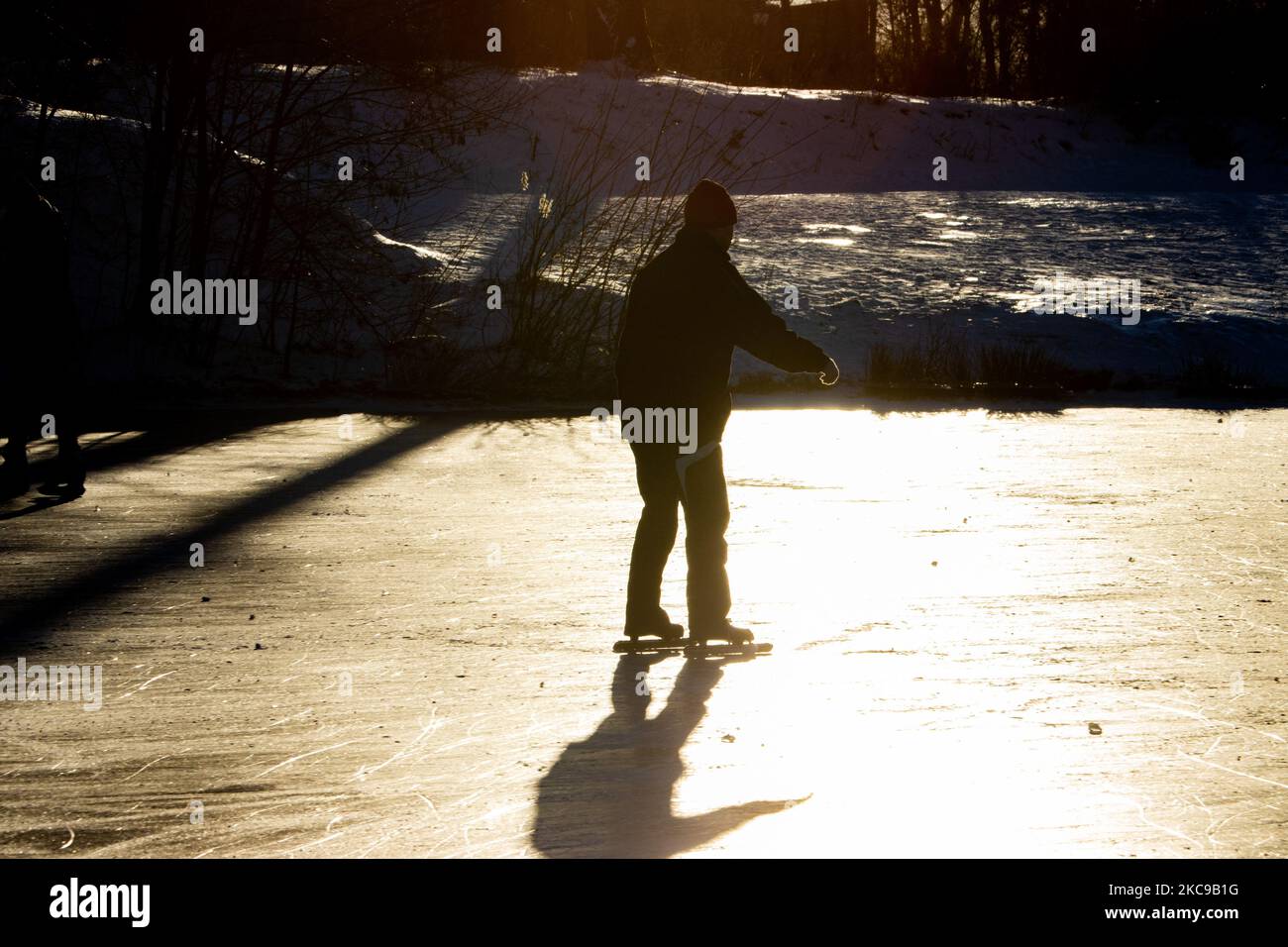 Dunkle Silhouetten von Menschen beim Eislaufen auf dem kristallklaren natürlichen gefrorenen See in der Region Nordbrabant am Nachmittag vor dem Sonnenuntergang. Eislaufrausch übernimmt die Niederlande. Die Niederländer haben viel Spaß und genießen das einwöchige, kalte Schneewetter mit Temperaturen unter Null, die dazu führen, dass Seen, Bäche, Teiche, Flüsse und Kanäle gefrieren. Im Park Meerland in der Nähe der Stadt Eindhoven haben sich die Massen an einem sonnigen Tag mit blauem Himmel abgesetzt, da es eine niederländische Tradition ist. Außerdem werden die Menschen im Freien in der eisgefrorenen Natur und der kalten Umgebung gesehen, die auf dem Eis der gefrorenen Seen spazieren gehen, p Stockfoto