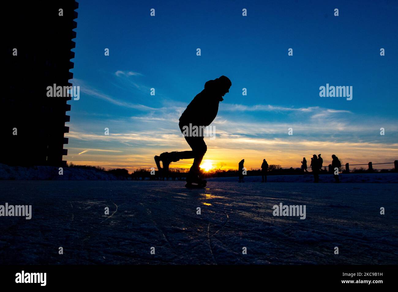 Dunkle Silhouetten von Menschen beim Eislaufen auf dem kristallklaren natürlichen gefrorenen See in der Region Nordbrabant am Nachmittag vor dem Sonnenuntergang. Eislaufrausch übernimmt die Niederlande. Die Niederländer haben viel Spaß und genießen das einwöchige, kalte Schneewetter mit Temperaturen unter Null, die dazu führen, dass Seen, Bäche, Teiche, Flüsse und Kanäle gefrieren. Im Park Meerland in der Nähe der Stadt Eindhoven haben sich die Massen an einem sonnigen Tag mit blauem Himmel abgesetzt, da es eine niederländische Tradition ist. Außerdem werden die Menschen im Freien in der eisgefrorenen Natur und der kalten Umgebung gesehen, die auf dem Eis der gefrorenen Seen spazieren gehen, p Stockfoto