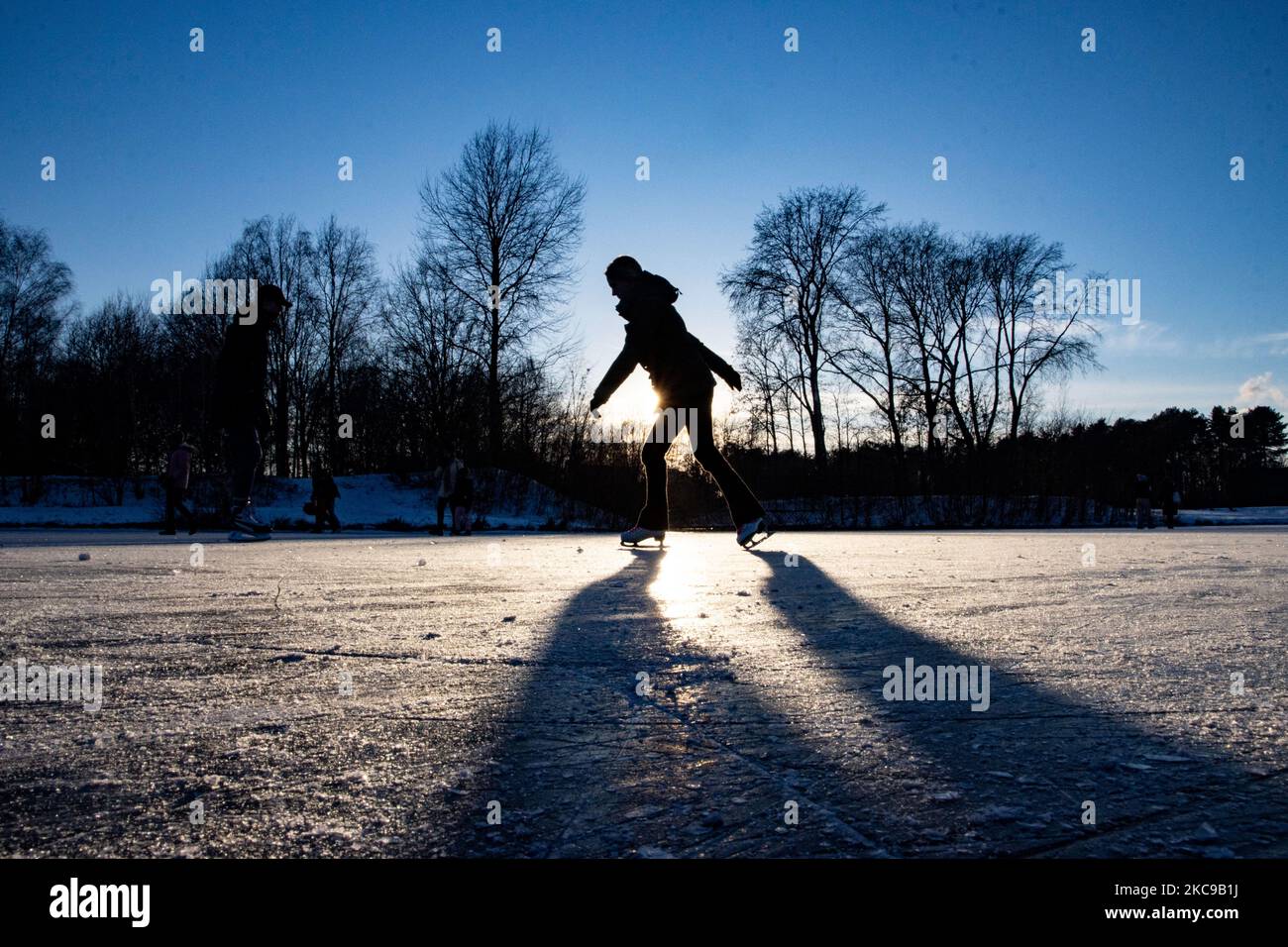 Dunkle Silhouetten von Menschen beim Eislaufen auf dem kristallklaren natürlichen gefrorenen See in der Region Nordbrabant am Nachmittag vor dem Sonnenuntergang. Eislaufrausch übernimmt die Niederlande. Die Niederländer haben viel Spaß und genießen das einwöchige, kalte Schneewetter mit Temperaturen unter Null, die dazu führen, dass Seen, Bäche, Teiche, Flüsse und Kanäle gefrieren. Im Park Meerland in der Nähe der Stadt Eindhoven haben sich die Massen an einem sonnigen Tag mit blauem Himmel abgesetzt, da es eine niederländische Tradition ist. Außerdem werden die Menschen im Freien in der eisgefrorenen Natur und der kalten Umgebung gesehen, die auf dem Eis der gefrorenen Seen spazieren gehen, p Stockfoto