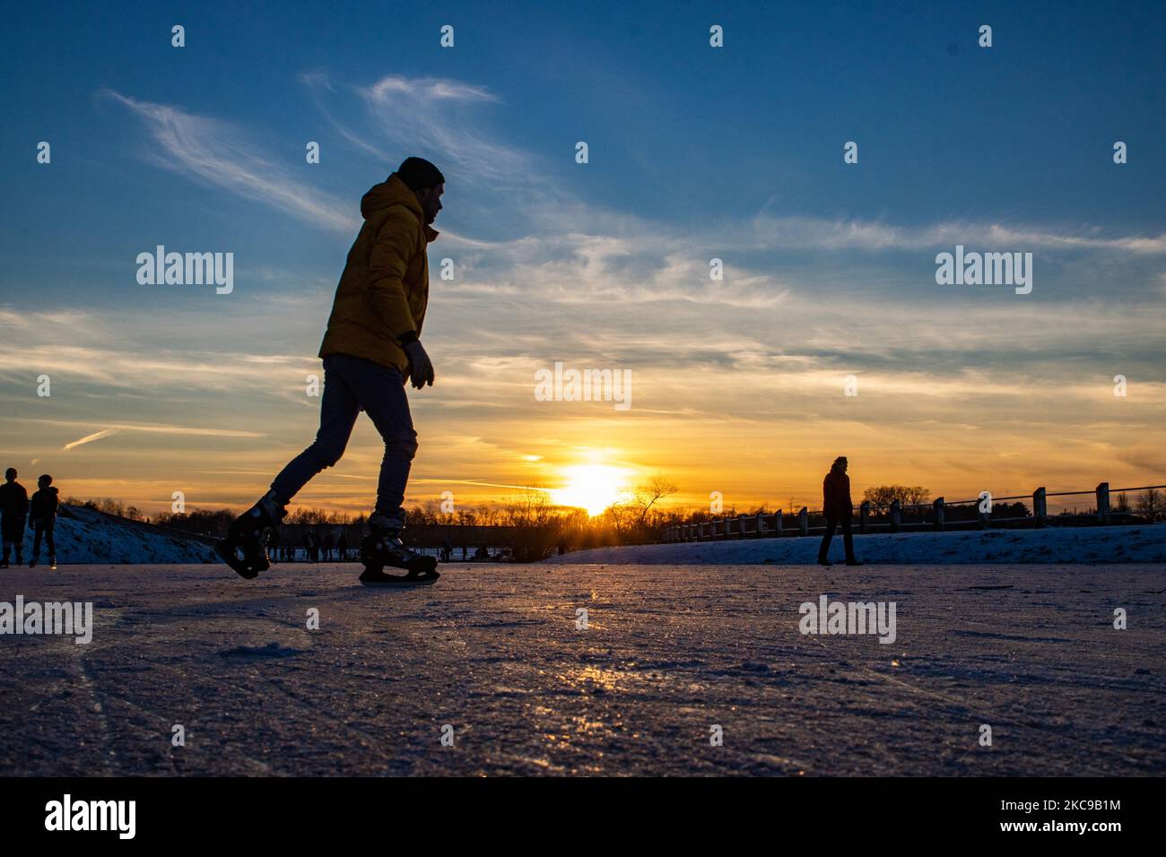 Dunkle Silhouetten von Menschen beim Eislaufen auf dem kristallklaren natürlichen gefrorenen See in der Region Nordbrabant am Nachmittag vor dem Sonnenuntergang. Eislaufrausch übernimmt die Niederlande. Die Niederländer haben viel Spaß und genießen das einwöchige, kalte Schneewetter mit Temperaturen unter Null, die dazu führen, dass Seen, Bäche, Teiche, Flüsse und Kanäle gefrieren. Im Park Meerland in der Nähe der Stadt Eindhoven haben sich die Massen an einem sonnigen Tag mit blauem Himmel abgesetzt, da es eine niederländische Tradition ist. Außerdem werden die Menschen im Freien in der eisgefrorenen Natur und der kalten Umgebung gesehen, die auf dem Eis der gefrorenen Seen spazieren gehen, p Stockfoto