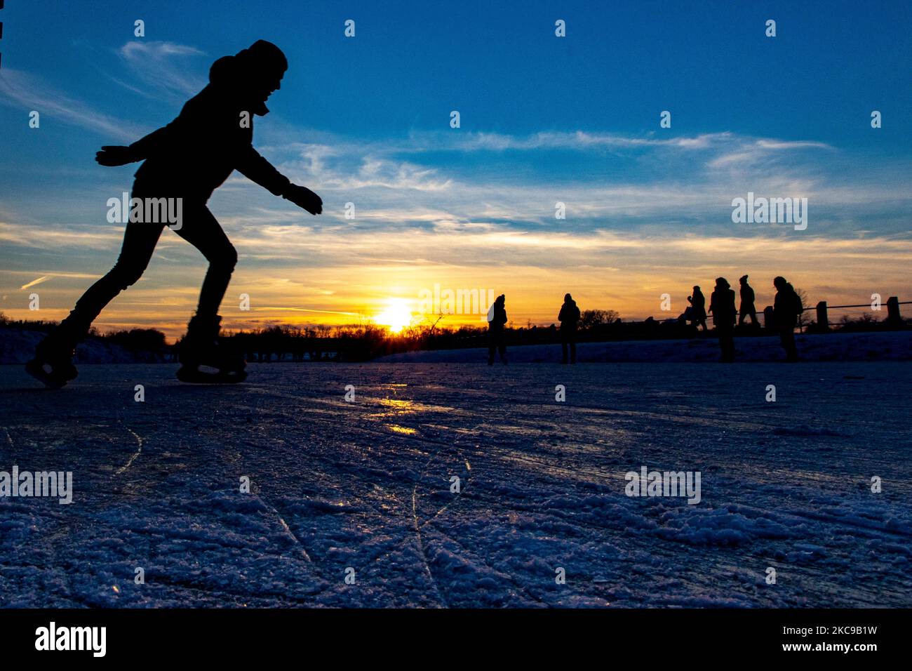 Dunkle Silhouetten von Menschen beim Eislaufen auf dem kristallklaren natürlichen gefrorenen See in der Region Nordbrabant am Nachmittag vor dem Sonnenuntergang. Eislaufrausch übernimmt die Niederlande. Die Niederländer haben viel Spaß und genießen das einwöchige, kalte Schneewetter mit Temperaturen unter Null, die dazu führen, dass Seen, Bäche, Teiche, Flüsse und Kanäle gefrieren. Im Park Meerland in der Nähe der Stadt Eindhoven haben sich die Massen an einem sonnigen Tag mit blauem Himmel abgesetzt, da es eine niederländische Tradition ist. Außerdem werden die Menschen im Freien in der eisgefrorenen Natur und der kalten Umgebung gesehen, die auf dem Eis der gefrorenen Seen spazieren gehen, p Stockfoto