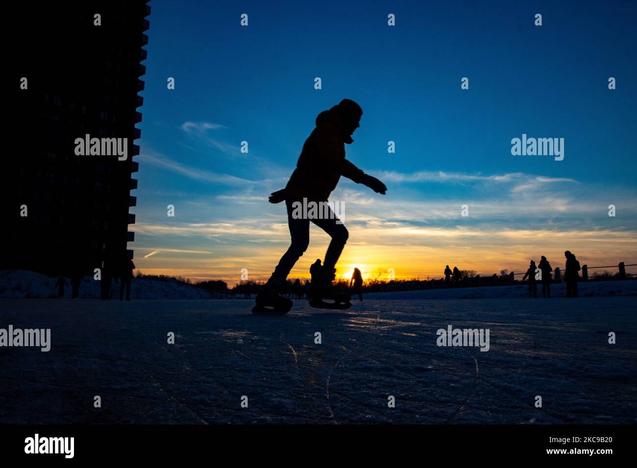 Dunkle Silhouetten von Menschen beim Eislaufen auf dem kristallklaren natürlichen gefrorenen See in der Region Nordbrabant am Nachmittag vor dem Sonnenuntergang. Eislaufrausch übernimmt die Niederlande. Die Niederländer haben viel Spaß und genießen das einwöchige, kalte Schneewetter mit Temperaturen unter Null, die dazu führen, dass Seen, Bäche, Teiche, Flüsse und Kanäle gefrieren. Im Park Meerland in der Nähe der Stadt Eindhoven haben sich die Massen an einem sonnigen Tag mit blauem Himmel abgesetzt, da es eine niederländische Tradition ist. Außerdem werden die Menschen im Freien in der eisgefrorenen Natur und der kalten Umgebung gesehen, die auf dem Eis der gefrorenen Seen spazieren gehen, p Stockfoto