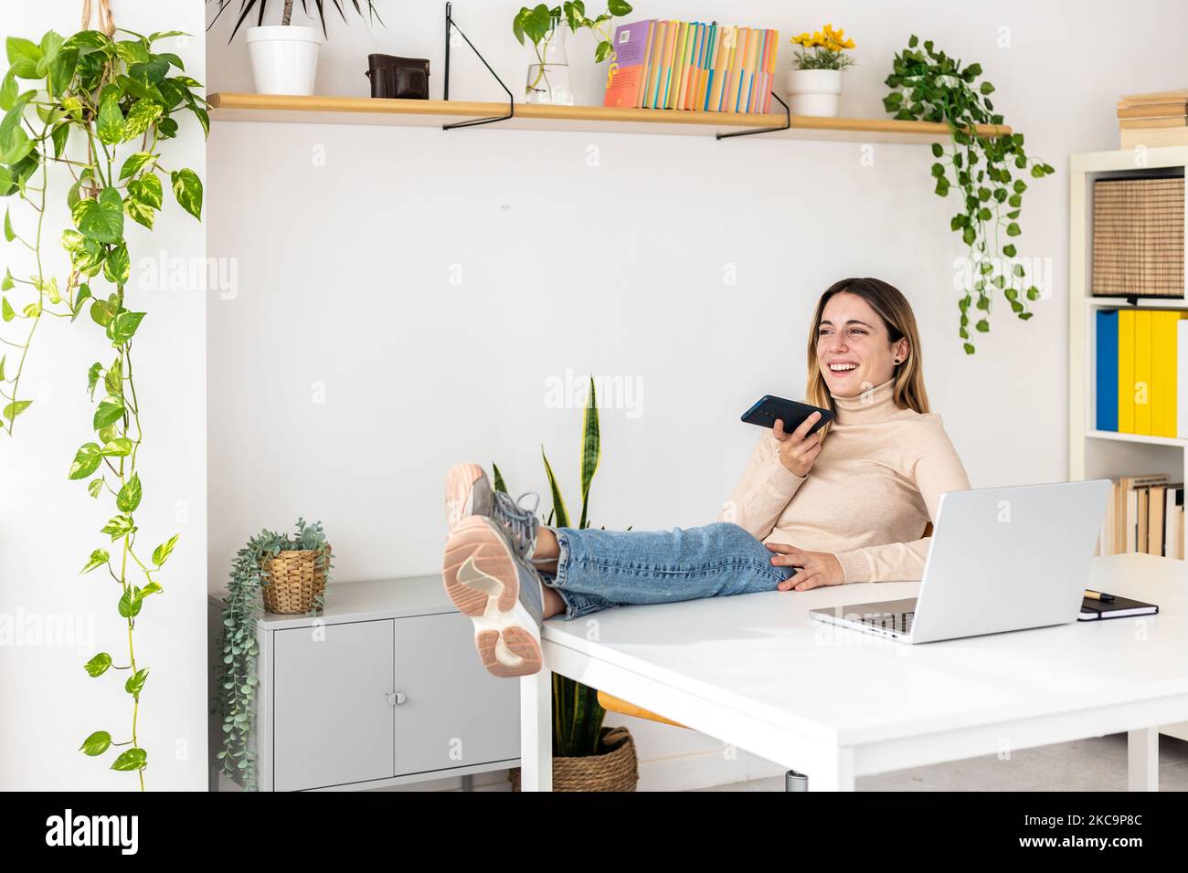 Junge Geschäftsfrau, die sich im Büro mit dem Mobiltelefon und den Füßen auf dem Schreibtisch entspannt Stockfoto