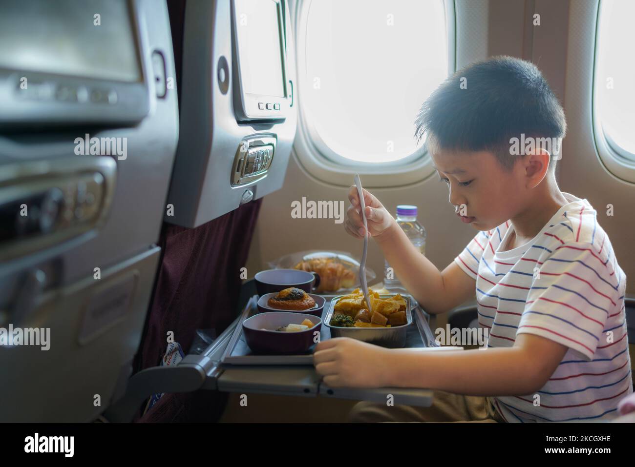Asiatisches Kind auf dem Fenstersitz des Flugzeugs. Kinderflugmahlzeit. Kinder fliegen. Spezielles Bordmenü, Speisen und Getränke für Baby und Kind. Junge, der gesundes Mittagessen isst Stockfoto