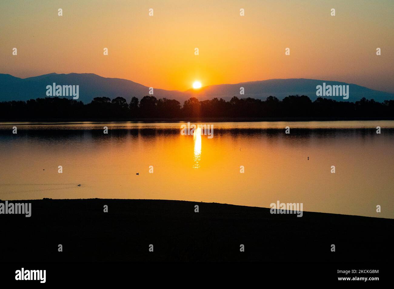 Die Sonne taucht hinter den Bergen auf und reflektiert sich auf der Wasseroberfläche. Magische Stunde Sommer Sonnenaufgang mit warmen Farben des Himmels und der Sonne über Kerkini mit Vögeln, Pferden, lokalen Fischern als Silhouetten gefischt, am See Nationalpark in Serres Region in Nordgriechenland. Der künstliche Kerkini-See ist ein einzigartiges Feuchtgebiet, ein Nationalpark und durch die Ramsar-Konvention als Feuchtgebiet mit Tausenden von Vögeln geschützt, darunter seltene und geschützte Flusswälder, da sich wichtige Hydrobiosphären entwickeln, die von großer internationaler Bedeutung und Akzeptanz sind, die wichtigste Wasserquelle Stockfoto