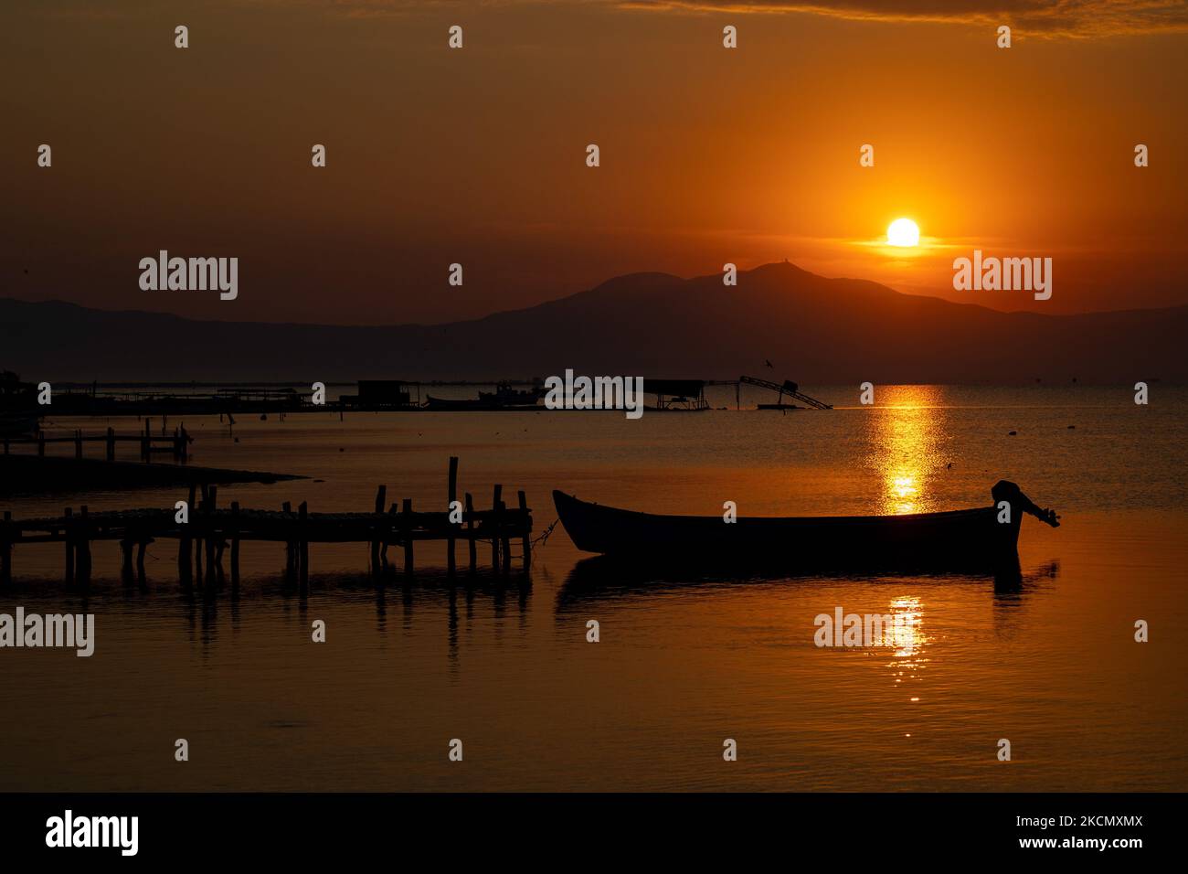 Die Sonne, die hinter dem Berg Hortiatis aufgeht, reflektiert das Wasser mit den Maschinen der Fischer und Muschelproduzenten, die in der Nähe der hölzernen Pfeiler sichtbar sind. Sonnenaufgang, Morgengrauen und die Sonne spiegeln sich auf dem Meer im Axios Delta National Park, einem Ort mit Feuchtgebieten in der Nähe von Halastra und Thessaloniki Stadt, wo 4 Flüsse Gallikos, Axios, Loudias und Aliakomnas ihr Delta haben. An dem jeweiligen Standort haben Fischer und Muschelproduzenten ihre Hütten und Boote. Über die Artenvielfalt ist diese Region berühmt und von weltweiter ornithologischer Bedeutung, da beträchtliche Populationen seltener Vögel anhalten, nisten oder überwinten Stockfoto