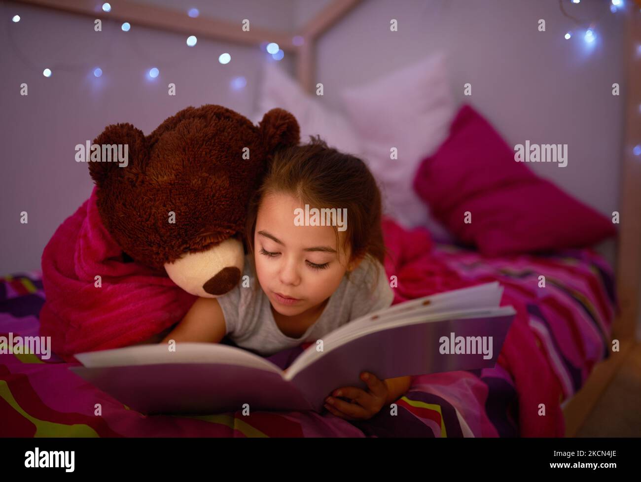 Es ist eine Geschichte, die die süßesten Träume entfachen soll. Ein kleines Mädchen, das mit ihrem Teddybär ein Buch im Bett liest. Stockfoto