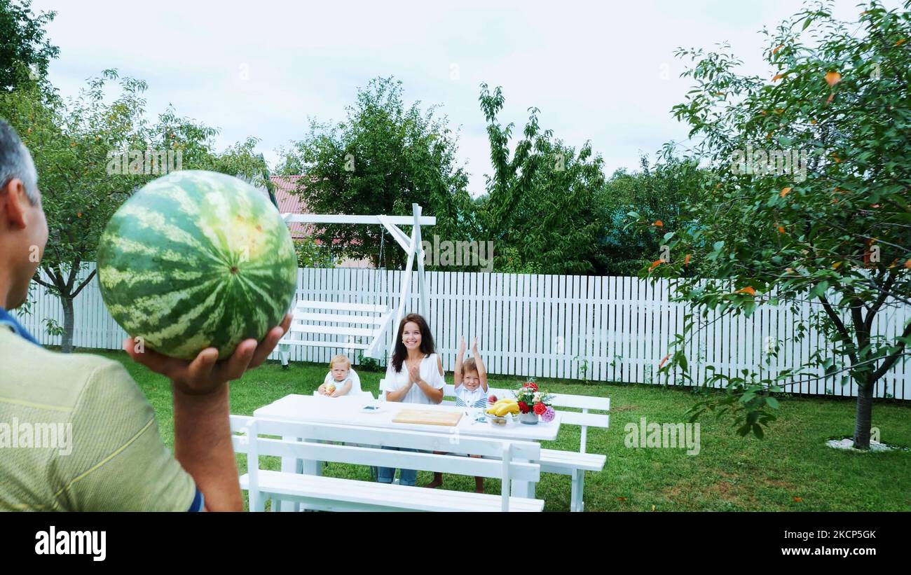 Sommer, im Garten, Vater trägt eine große Wassermelone, wird seine Familie zu behandeln. Mittagessen mit der Familie. Die Familie verbringt ihre Freizeit miteinander. Hochwertige Fotos Stockfoto