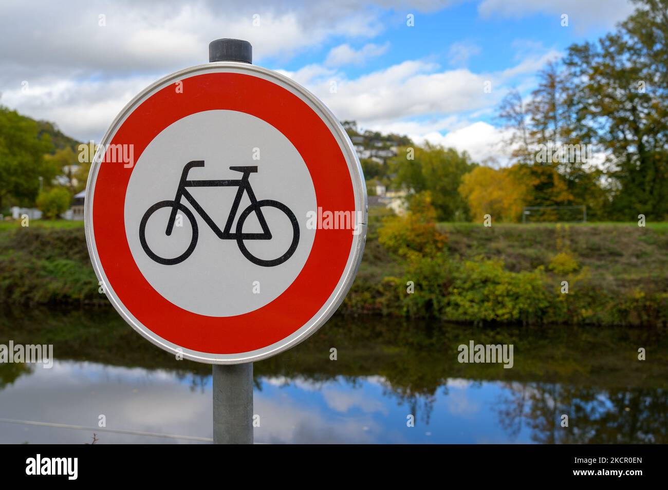 Fahrräder verboten oder kein Fahrradschild vor einem kleinen Fluss und fallen farbige Bäume mit blauem Himmel und weißen geschwollenen Wolken, die sich im Wasser spiegeln Stockfoto