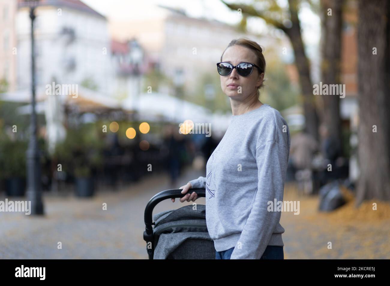 Eine stilvolle junge Frau mit Sonnenbrille, die durch die Innenstadt von Ljubljana geht und dabei den Kinderwagen schob und schaukelte. Warmes Herbst- oder Frühlingswetter für Outdoor-Aktivitäten Stockfoto