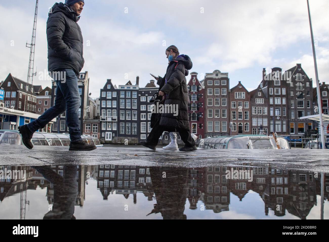 Menschen mit ihren Reflexionen, die vor den liegenden Häusern von Amsterdam spazieren. Einheimische und ein paar Touristen in den ruhigen Straßen von Amsterdam während der Sperre in der niederländischen Hauptstadt mit Geschäften und Geschäften, die mit geschlossenen Rollläden aus Metall erscheinen, Cafés, Bars und Restaurants auch geschlossen mit Tischen und Stühlen der Terrassen verschlossen. Die Niederlande waren die erste europäische Nation, die die vollständige Sperre erklärte, um gegen die neue Omicron-Variante zu kämpfen, die aufkommt. Nach einem plötzlichen Regierungsbefehl vor Weihnachten schloss das Land alle unwesentlichen Geschäfte, Cafés, Restaurants, Bars und Fitnessstudios Stockfoto