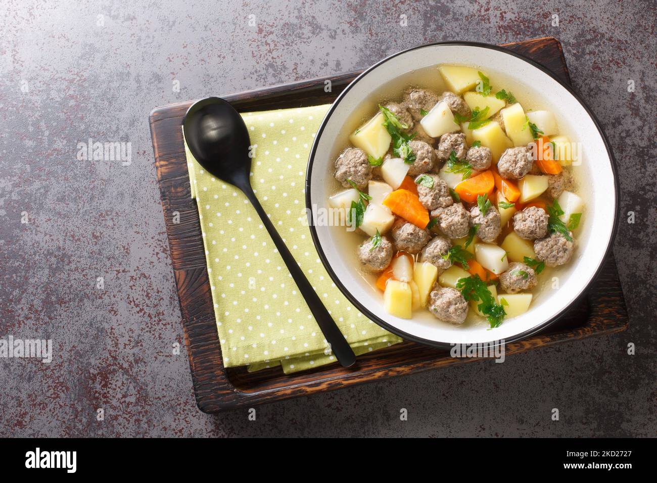 Köstliche Suppe mit Wurzelgemüse und Fleischbällchen in einer klaren Brühe in Nahaufnahme auf einem Teller auf dem Tisch. Horizontale Draufsicht von oben Stockfoto
