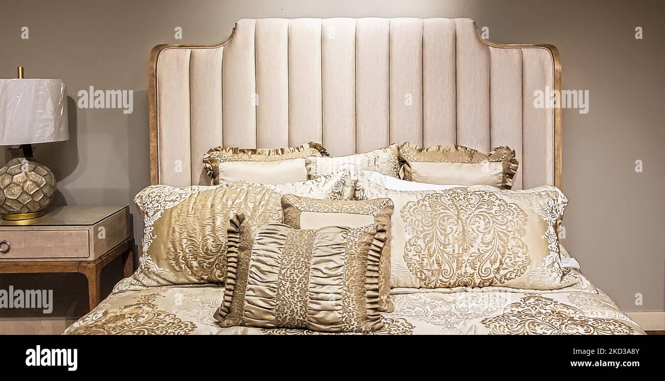 Eleganter, neutraler Satin-Bettbezug und Kissen am gepolsterten Bettgestell und an der beigefarbenen Wand mit passender Lampe Stockfoto
