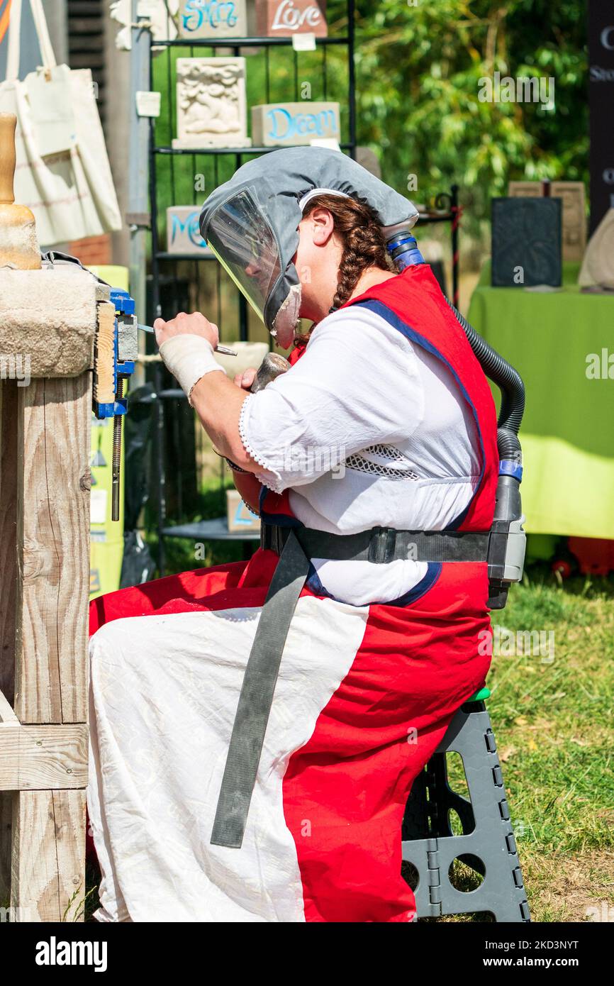 Frau trägt eine schützende Gesichtsmaske mit Luftzufuhr, während sie ein Stück Stein mit Meißel und Hammer im Freien bei einer Veranstaltung modelliert und graviert. Stockfoto