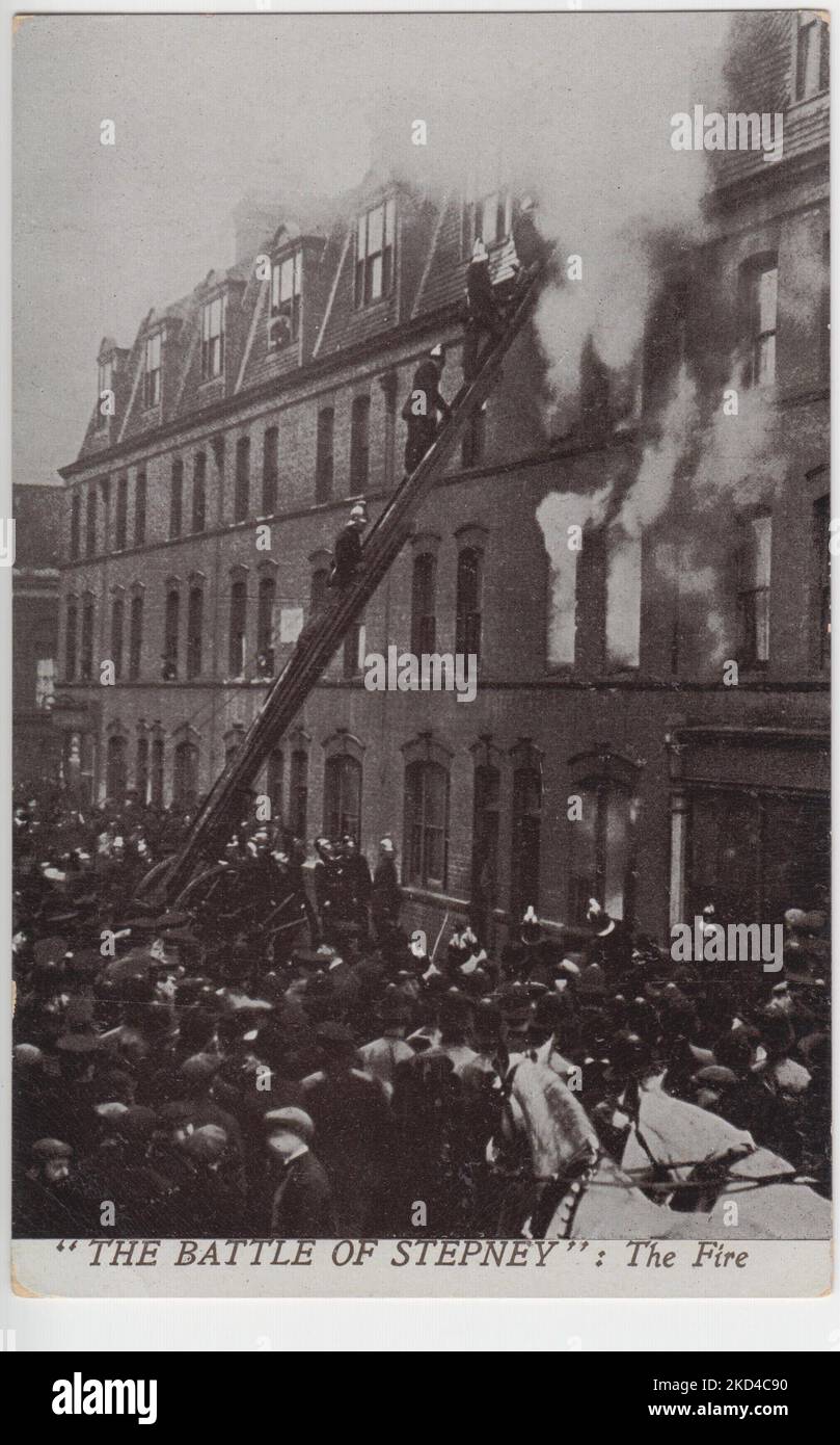 „The Battle of Stepney: The Fire“: Postkarte der Belagerung der Sidney Street von 1911, zeigt die Sidney Street von 100 in Flammen, wobei Rauch aus den Fenstern des oberen Stockwerks strömt. Feuerwehrleute stehen auf einer Leiter, die sich gegen das Gebäude lehnt, eine große Gruppe von Zuschauern ist draußen Stockfoto