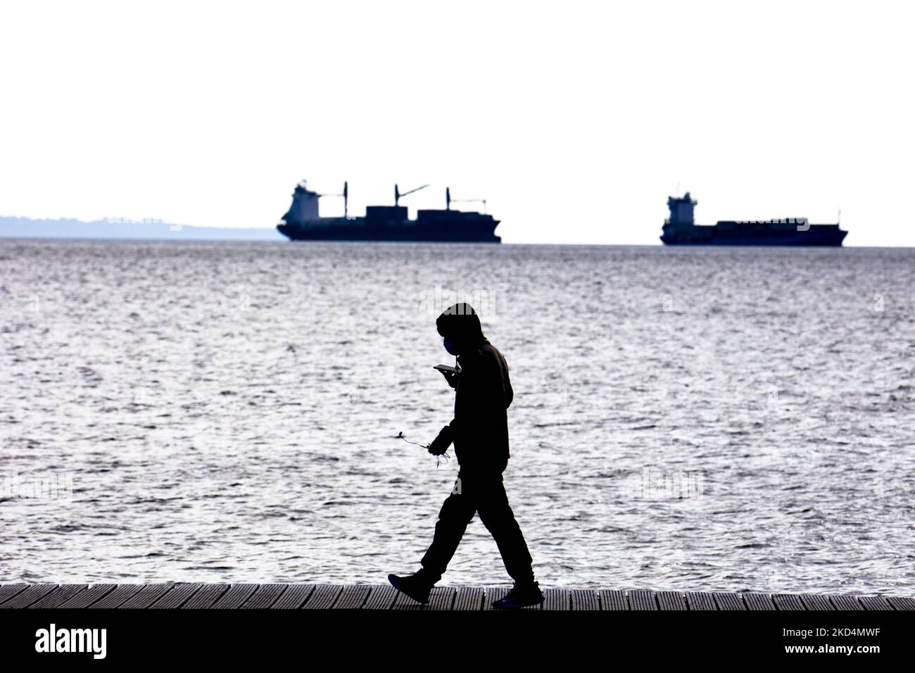 Silhouette eines Mannes, der sein Telefon mit Frachtschiffen im Hintergrund hält. Tägliches Leben in Thessaloniki, der zweitgrößten Stadt Griechenlands. Die lange Uferpromenade der Stadt ist ein Wahrzeichen und Anziehungspunkt für Einheimische und Touristen, da die Stadt städtische Umgebung, traditionelle und historische Stätten mit zeitgenössischen, wunderschönen Meer- und Bergketten verbindet. Griechenland hat immer noch strenge Maßnahmen gegen die Coronavirus-Pandemie Covid-19, wie die obligatorische Verwendung von medizinischer Gesichtsmask im Freien, in Thessaloniki, Griechenland am 5. März 202 Stockfoto