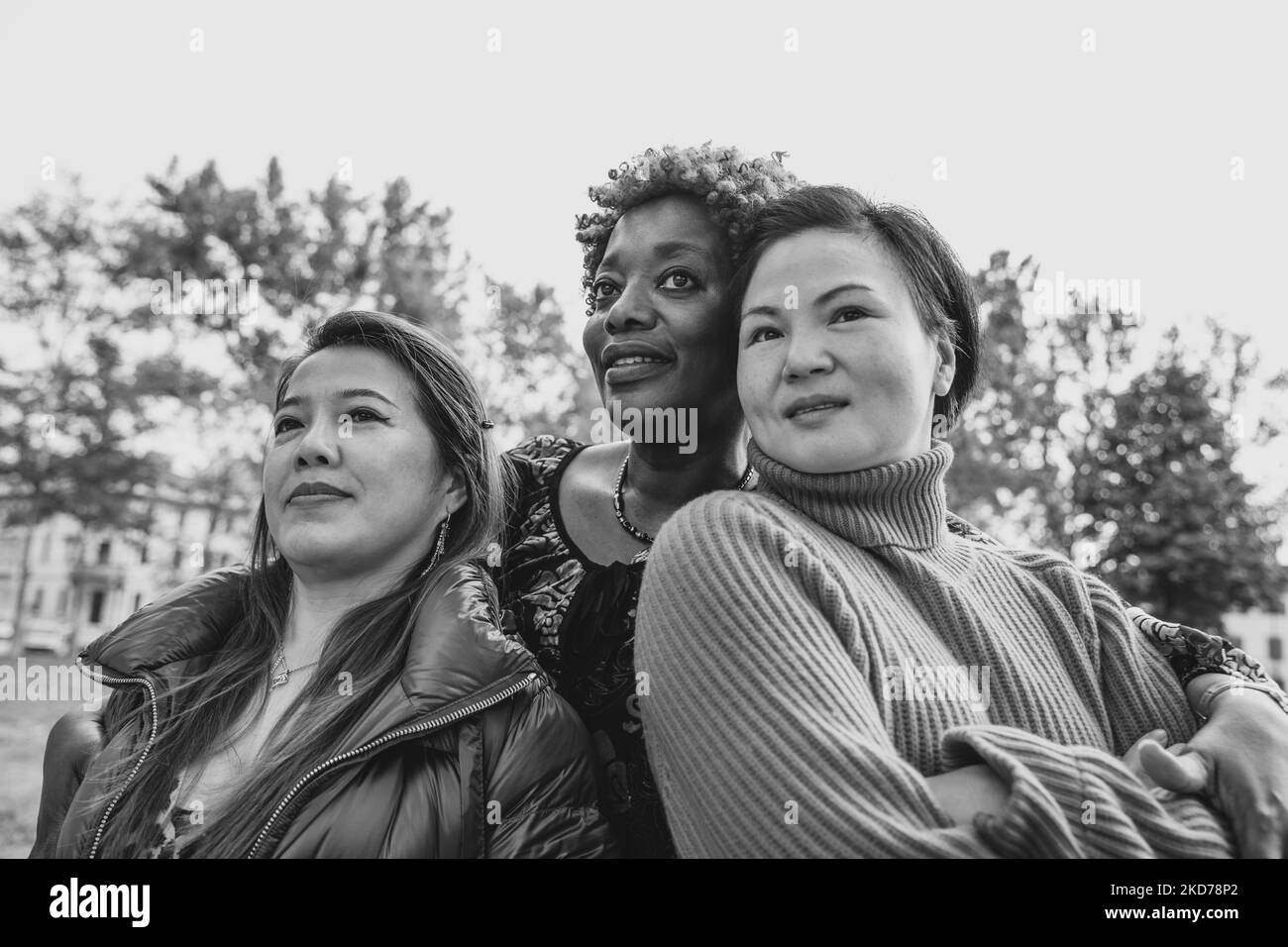 Schwarzweiß-Foto von drei multirassischen Freunden, die sich im Stadtpark umarmen und nach oben schauen. Stockfoto