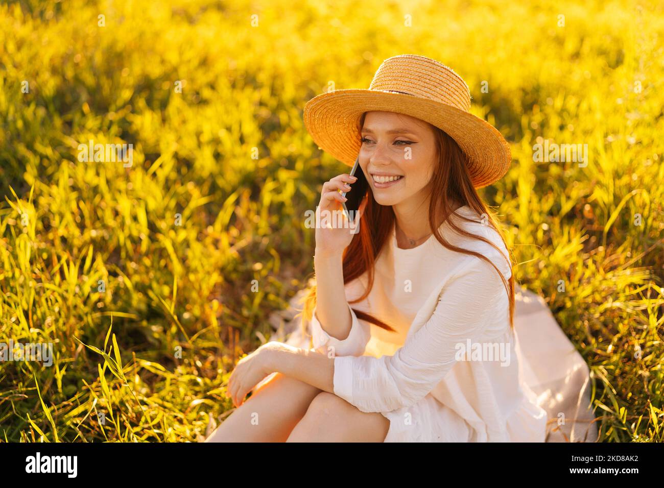 Mittellange Porträtaufnahme einer charmanten, positiven rothaarigen jungen Frau mit Strohhut und weißem Kleid, die auf dem Smartphone auf einem schönen Feld sitzt. Stockfoto