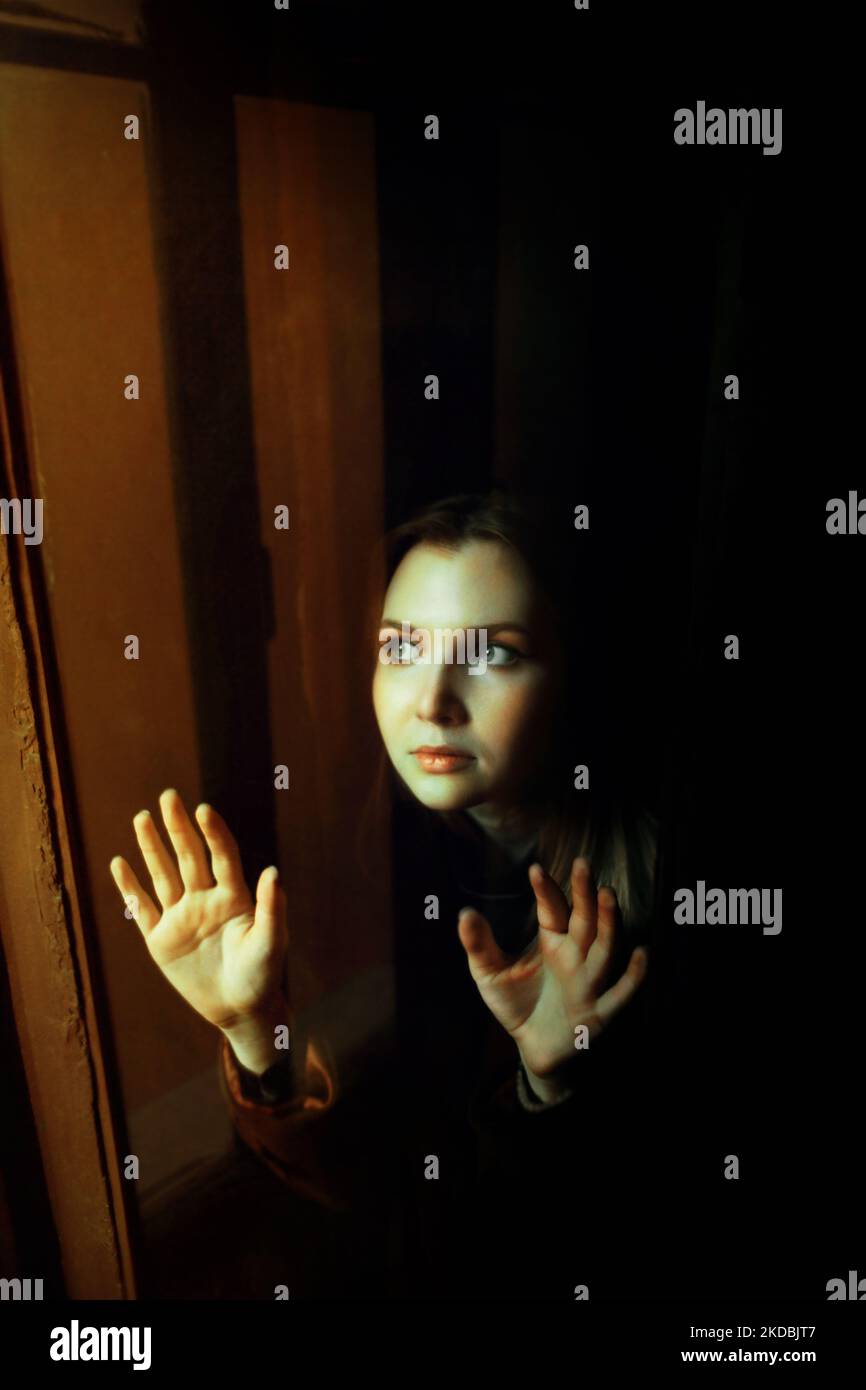 Porträt einer schönen blonden Frau, die in der Abenddämmerung mit ihren Händen die Glasoberfläche eines offenen Fensters berührt. Mystery. Stockfoto