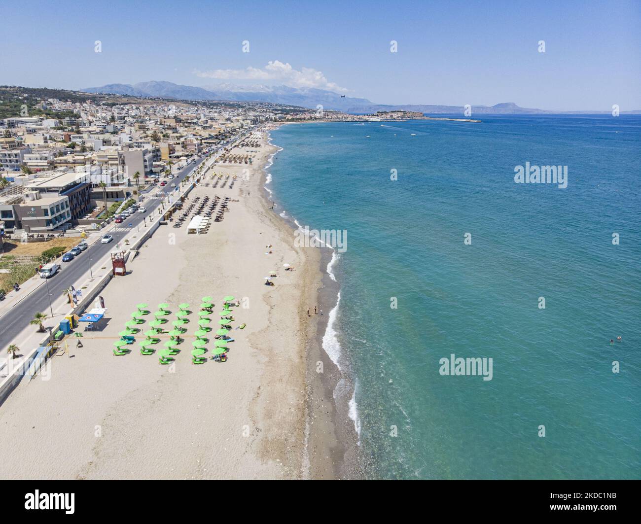 Luftaufnahmen von einer Drohne der Küste der Stadt Rethymno mit dem langen Strand und den Strandbars auf der Insel Creta. Man sieht die Menschen an der Strandbar unter dem Sonnenschirm die Sonne genießen oder im kristallklaren Meer schwimmen. Rethymno ist eine historische mediterrane Strandstadt an der Nordküste Kretas, die an der Ägäis liegt und 40,000 Einwohner zählt. Ein touristisches Ziel mit einem historischen venezianischen Hafen und einer Stadt, archäologischen Stätten, endlosen Sandstränden, Wassersportmöglichkeiten, schönen, traditionellen Tavernen und einer großen Auswahl an Hotels. Der Tourismus kehrt zurück, und die Vorbehalte steigen Stockfoto