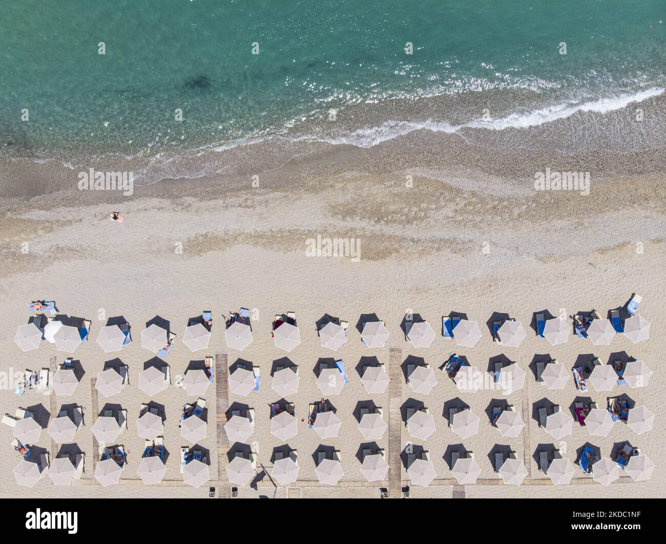 Luftaufnahmen von einer Drohne der Küste der Stadt Rethymno mit dem langen Strand und den Strandbars auf der Insel Creta. Man sieht die Menschen an der Strandbar unter dem Sonnenschirm die Sonne genießen oder im kristallklaren Meer schwimmen. Rethymno ist eine historische mediterrane Strandstadt an der Nordküste Kretas, die an der Ägäis liegt und 40,000 Einwohner zählt. Ein touristisches Ziel mit einem historischen venezianischen Hafen und einer Stadt, archäologischen Stätten, endlosen Sandstränden, Wassersportmöglichkeiten, schönen, traditionellen Tavernen und einer großen Auswahl an Hotels. Der Tourismus kehrt zurück, und die Vorbehalte steigen Stockfoto