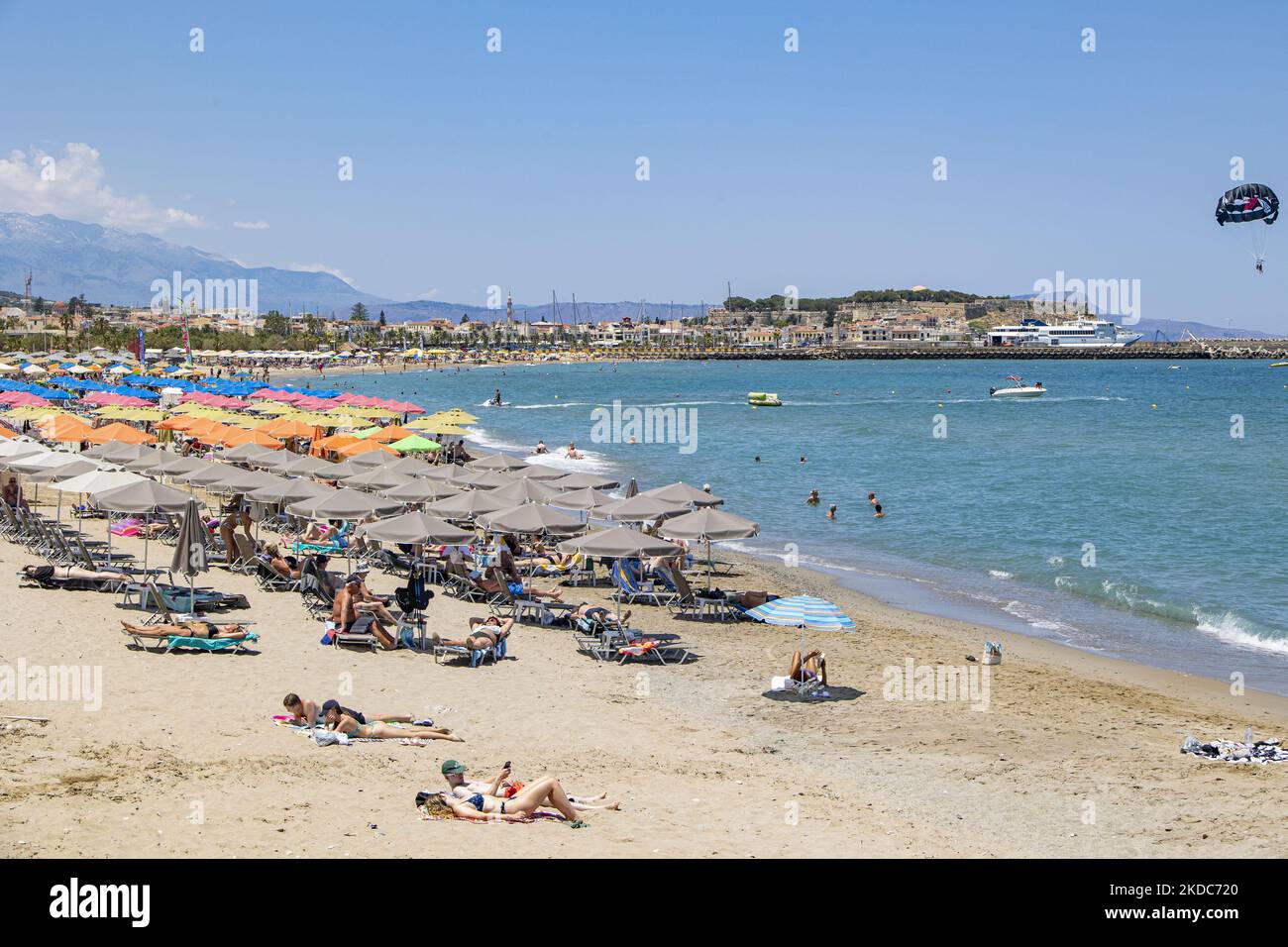 Tourismus in Griechenland. Die Küste der Stadt Rethymno mit dem langen Sandstrand und den Strandbars auf der Insel Creta mit den Bergen im Hintergrund. Man sieht die Menschen beim Sonnenbaden in Badeanzug, Badeanzug, Bikini, beim Sonnenbaden unter dem Sonnenschirm an der Strandbar auf einer Sonnenliege und beim Schwimmen im kristallklaren Meer. Rethymno ist eine historische mediterrane Strandstadt an der Nordküste Kretas, die an der Ägäis liegt und 40,000 Einwohner zählt. Ein touristisches Ziel mit einem historischen venezianischen Hafen und einer Stadt, archäologischen Stätten, endlosen Sandstränden, Wassersportmöglichkeiten, schönen schönen T Stockfoto