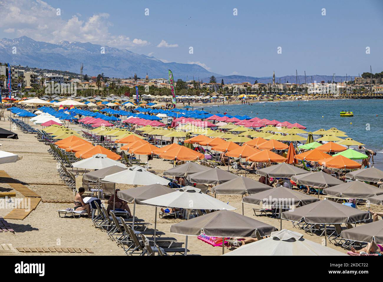 Tourismus in Griechenland. Die Küste der Stadt Rethymno mit dem langen Sandstrand und den Strandbars auf der Insel Creta mit den Bergen im Hintergrund. Man sieht die Menschen beim Sonnenbaden in Badeanzug, Badeanzug, Bikini, beim Sonnenbaden unter dem Sonnenschirm an der Strandbar auf einer Sonnenliege und beim Schwimmen im kristallklaren Meer. Rethymno ist eine historische mediterrane Strandstadt an der Nordküste Kretas, die an der Ägäis liegt und 40,000 Einwohner zählt. Ein touristisches Ziel mit einem historischen venezianischen Hafen und einer Stadt, archäologischen Stätten, endlosen Sandstränden, Wassersportmöglichkeiten, schönen schönen T Stockfoto