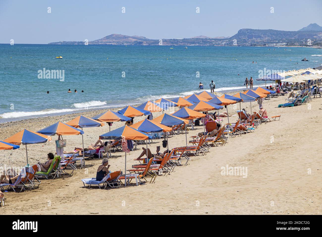 Tourismus in Griechenland. Die Küste der Stadt Rethymno mit dem langen Sandstrand und den Strandbars auf der Insel Creta mit den Bergen im Hintergrund. Man sieht die Menschen beim Sonnenbaden in Badeanzug, Badeanzug, Bikini, beim Sonnenbaden unter dem Sonnenschirm an der Strandbar auf einer Sonnenliege und beim Schwimmen im kristallklaren Meer. Rethymno ist eine historische mediterrane Strandstadt an der Nordküste Kretas, die an der Ägäis liegt und 40,000 Einwohner zählt. Ein touristisches Ziel mit einem historischen venezianischen Hafen und einer Stadt, archäologischen Stätten, endlosen Sandstränden, Wassersportmöglichkeiten, schönen schönen T Stockfoto