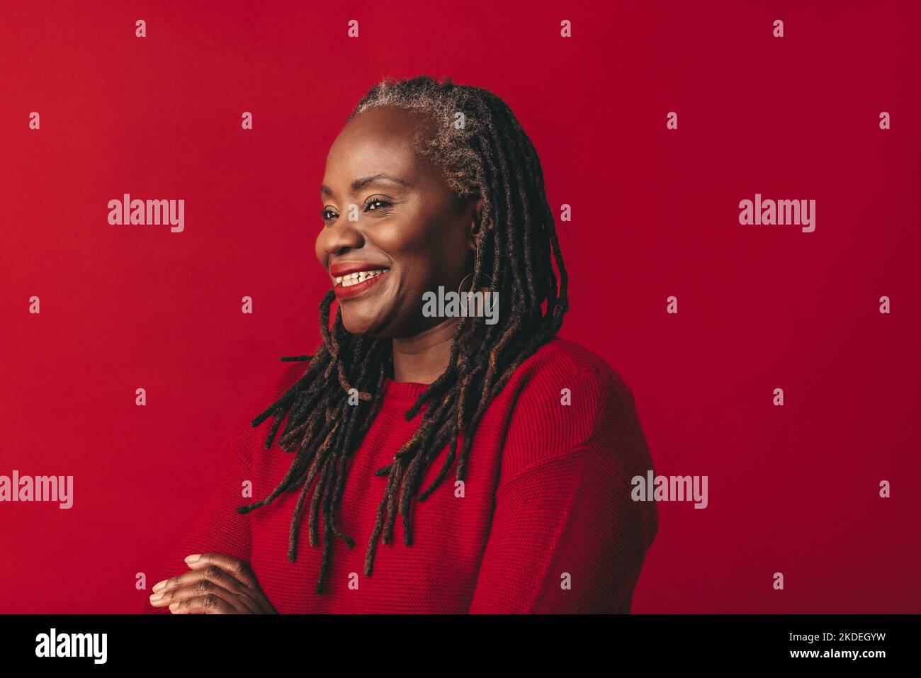 Fröhliche schwarze Frau lächelt und schaut weg, während sie vor einem roten Hintergrund steht. Glückliche reife Frau umarmt ihr natürliches Haar mit Selbstvertrauen. Stockfoto