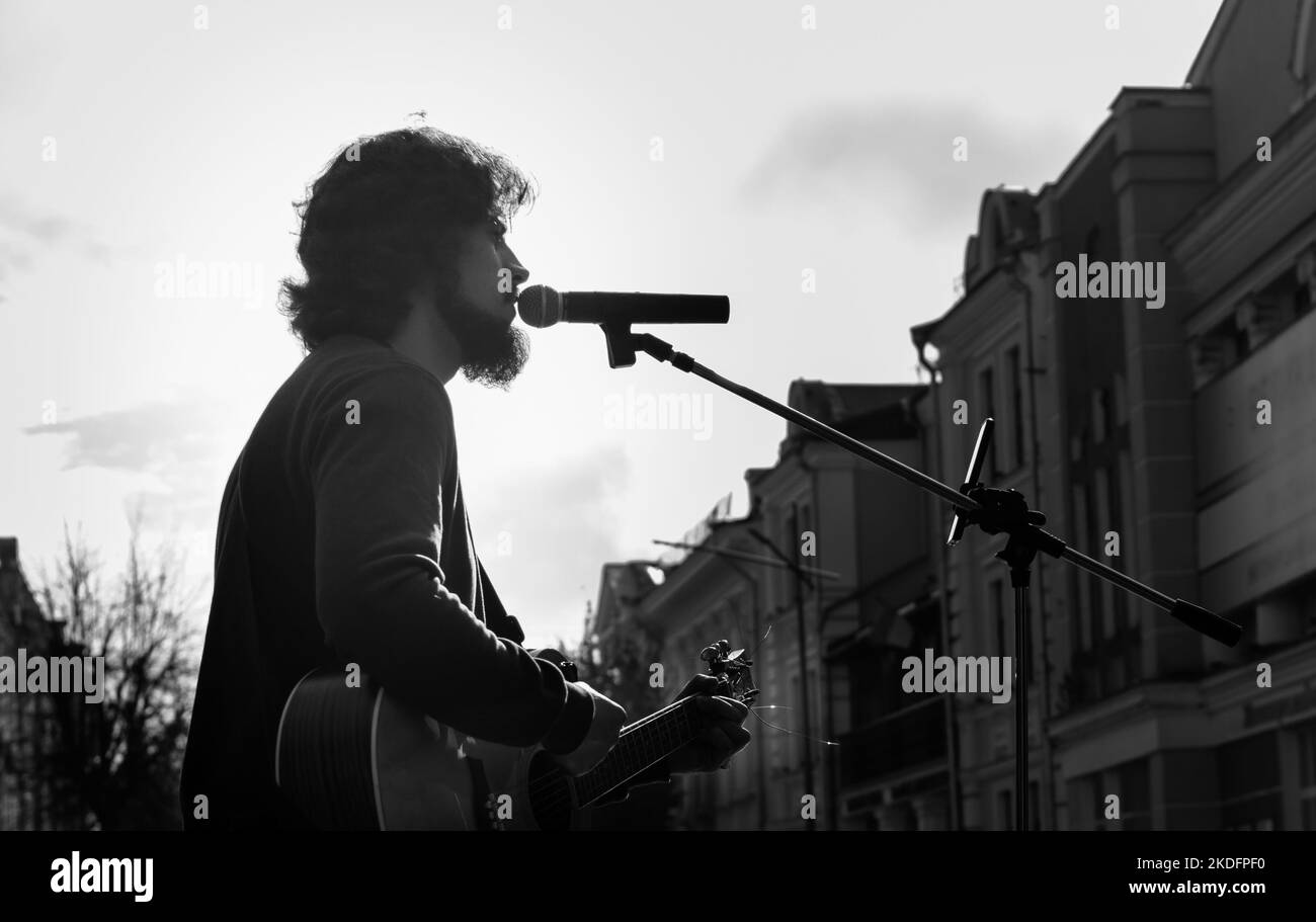 Straßenmusiker, der im Mikrofon auf unscharfem Hintergrund von Stadtstraßengebäuden und grauem Himmel singt. Ukraine, Zhytomyr, 04. November 2022 Stockfoto