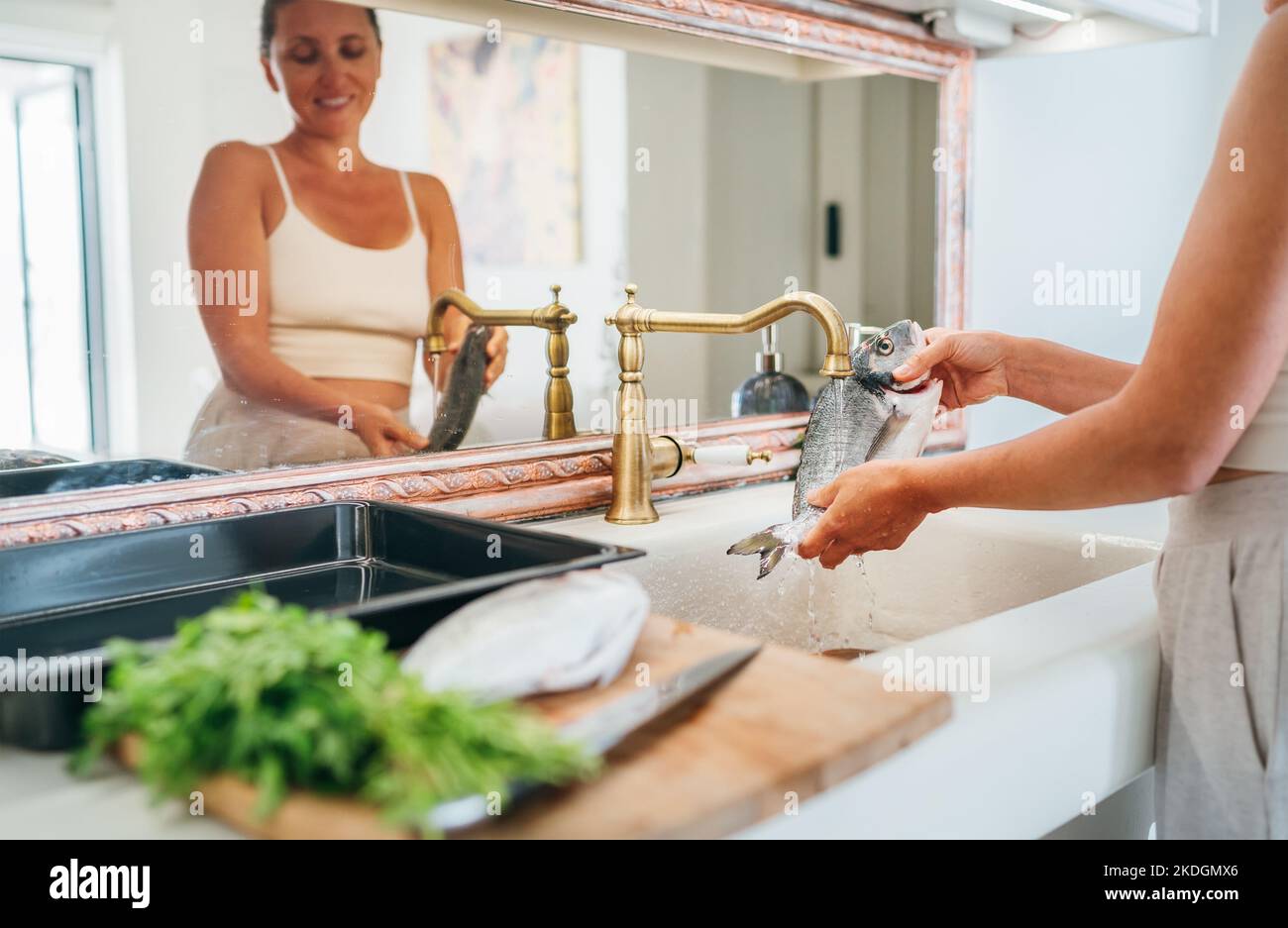 Lächelnde junge Erwachsene Frau, die Fische unter dem Wasserhahn der Küchenspüle waschen. Gesunde Meeresfrüchte Zubereitung und Hausarbeit Konzept Bild. Stockfoto
